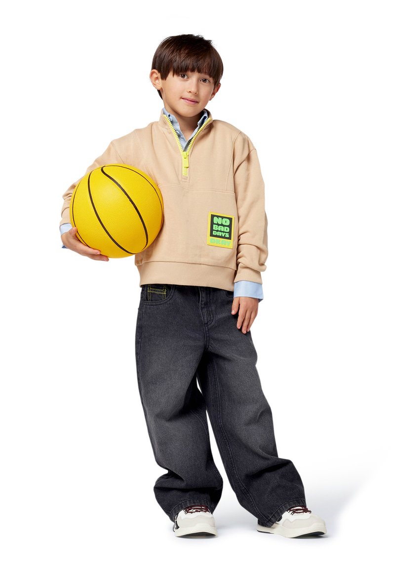Junger Junge hält einen gelben Basketball, trägt einen beigen Sweatshirt mit dem Aufnäher "NO BAD DAYS", schwarze baggy Jeans und weiße Sneakers vor einem weißen Hintergrund.