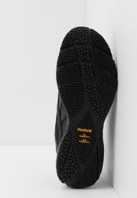 Suela de zapatilla Reebok negra con goma texturizada, que presenta patrones circulares y en zigzag, etiquetada como resistente al aceite y deslizamiento en amarillo.