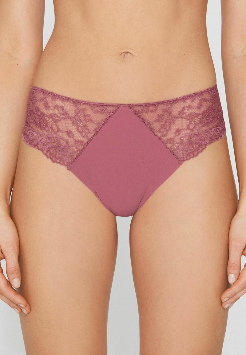 Mujer con ropa interior estilo bikini en encaje malva y tela sólida, mostrada desde la cintura hasta la mitad del muslo, con las manos relajadas a los lados.