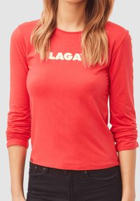 Camiseta de manga larga roja hecha de algodón, con un escote redondo y la palabra "LAGAT" impresa en letras blancas en negrita en el pecho.