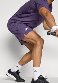 Lila träningsskorta och shorts med en slät textur. Svarta sportskor med vita detaljer. Vita ankelsockar med logotyp.