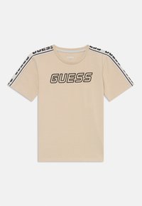 Beige katoenen T-shirt met korte mouwen, voorzien van een groot zwart 'GUESS' logo en contrasterende zwart-witte strepen op de schouders.