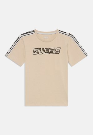 Bežīgs kokvilnas t-krekls ar īsām piedurknēm, kurā redzams liels melns "GUESS" logotips un kontrastējošas melnas un baltas svītras uz pleciem.