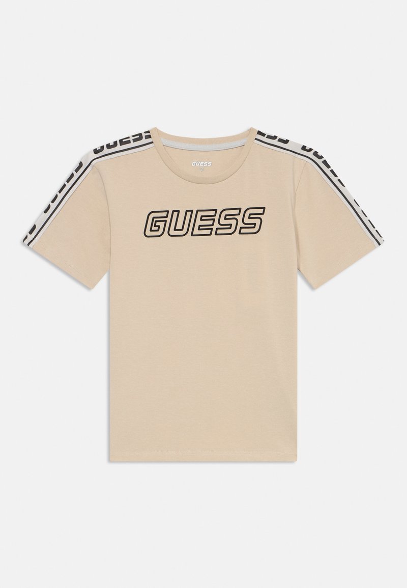 Beige katoenen T-shirt met korte mouwen, voorzien van een groot zwart 'GUESS' logo en contrasterende zwart-witte strepen op de schouders.