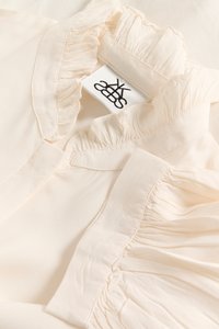 Blouse couleur crème avec col et manches volantés, fabriquée en tissu léger, avec une étiquette cousue affichant un logo.