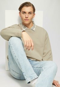 Felpa beige con polsini a costine sopra una camicia a quadri, abbinata a jeans azzurri chiari. Texture liscia, vestibilità casual. Sfondo neutro.