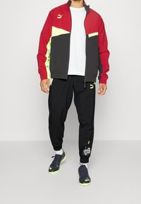 Puma Sportjacke in Rot, Schwarz und Neon-Gelb, kombiniert mit schwarzen Jogginghosen mit Akzenten und Branding, getragen mit schwarzen Schuhen.