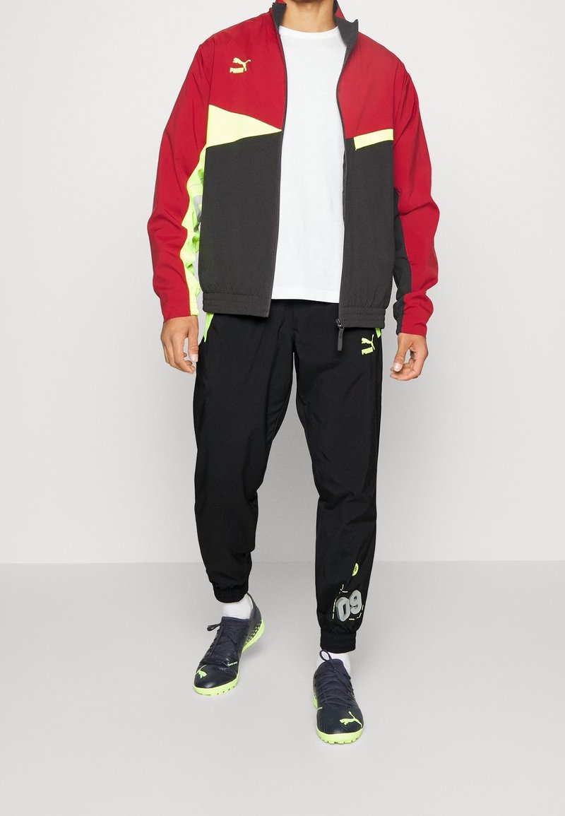 Puma Sportjacke in Rot, Schwarz und Neon-Gelb, kombiniert mit schwarzen Jogginghosen mit Akzenten und Branding, getragen mit schwarzen Schuhen.