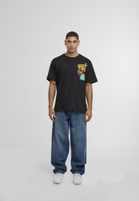 Fekete pamut póló grafikai foltokkal piros, sárga és zöld színben; laza kék farmernadrággal és fehér sportcipőkkel párosítva.