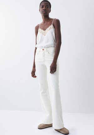 Jeans acampanados blancos combinados con una camiseta de seda ligera con un dobladillo de encaje en el escote. La modelo está de pie frente a un fondo neutro.