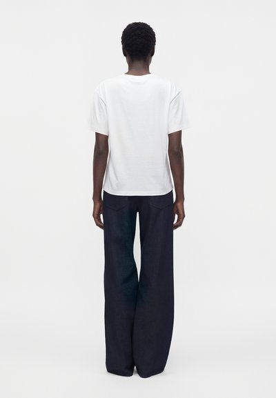 Person standing facing away, wearing a loose white short-sleeve shirt and wide-leg dark trousers against a plain background.