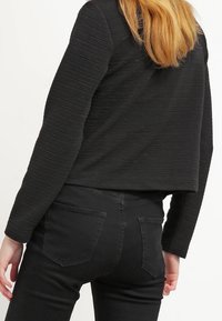 Černý žebrovaný crop top s dlouhými rukávy, zdobený texturovaným vzorem. Spárován s černými džíny s kapsami na zadní straně a přiléhavým střihem.