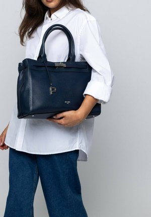 Woman holding a large navy blue leather handbag with silver hardware, wearing a white button-up shirt and blue jeans.