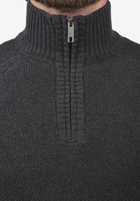 Grauer Strickpullover mit hohem Kragen und halbem Reißverschluss. Die Struktur ist gerippt, mit einem markanten Muster im gesamten Stoff.