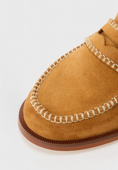Mocasines de ante en color tostado, con costuras en color crema a lo largo de los bordes, puntera redondeada y suela de goma con un fino ribete.