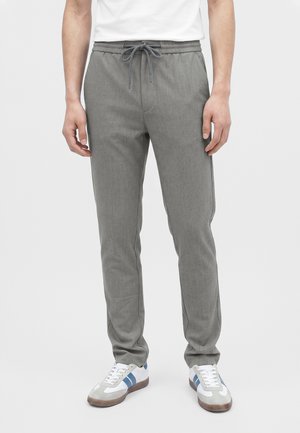 Mand iført grå snøre bukser og hvide sneakers med blå striber, står mod en ensfarvet lys baggrund.