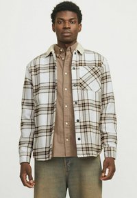 Geruite flanellen shirt in wit, bruin en zwart met een beige kraag, voork pocket en drukknoopsluitingen, gedragen over een bruine button-up shirt.