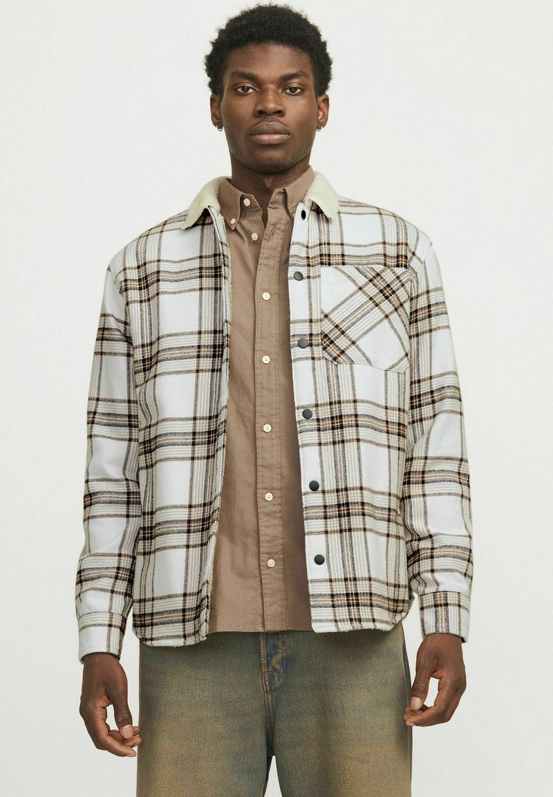 Geruite flanellen shirt in wit, bruin en zwart met een beige kraag, voork pocket en drukknoopsluitingen, gedragen over een bruine button-up shirt.