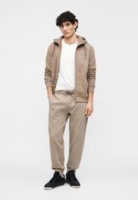 Beige hoodie met rits over een wit T-shirt, gecombineerd met beige joggers en zwarte sneakers. Gladde stof en ontspannen pasvorm.