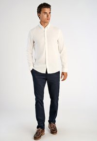 Camicia bianca a maniche lunghe con bottoni e finitura testurizzata, abbinata a pantaloni blu navy scuro e scarpe da barca marroni con lacci.