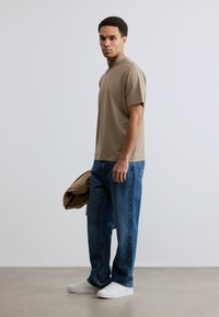 Ανοιχτό καφέ βαμβακερό μπλουζάκι με ψηλό λαιμό, συνδυασμένο με μπλε τζιν και λευκά αθλητικά παπούτσια, κρατώντας ένα μπεζ μπουφάν πάνω σε ένα χέρι.