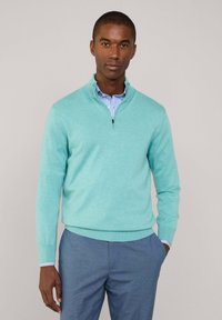 Maglione blu chiaro con zip in tessuto morbido e finitura testurizzata, abbinato a una camicia a righe con colletto e pantaloni blu.