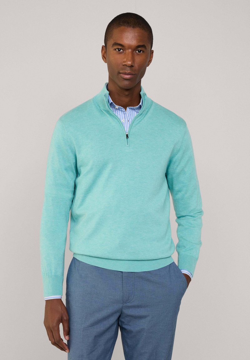 Maglione blu chiaro con zip in tessuto morbido e finitura testurizzata, abbinato a una camicia a righe con colletto e pantaloni blu.