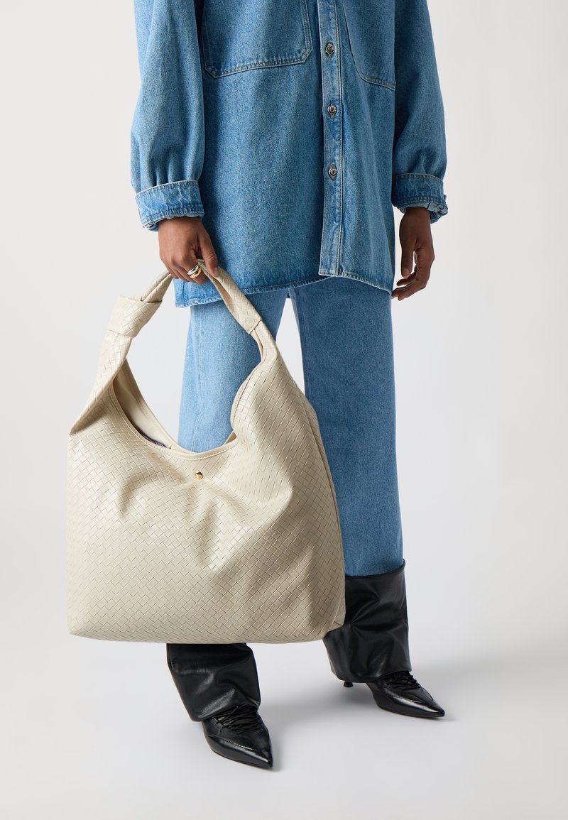 Sac en tissu tissé beige avec une grande forme décontractée, doté d'une poignée arrondie et d'un subtil accent doré, porté par une personne en tenue en denim.