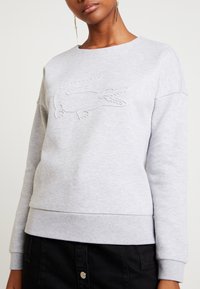 Femme portant un sweat-shirt gris clair avec un logo crocodile en relief et une jupe noire à boutons, debout devant un fond uni.