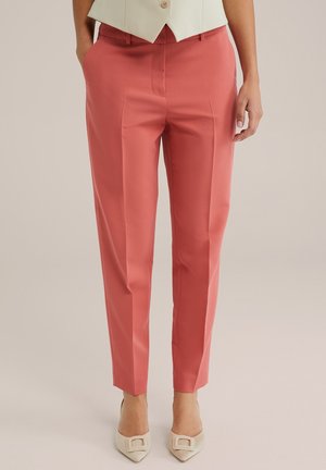 Mujer con pantalones hasta el tobillo personalizados color coral con pliegue al frente, manos en los bolsillos, combinados con tacones crema de punta afilada.