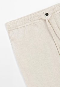 Beige linen pants with a drawstring waist, featuring a brown button, soft texture, and a streamlined design.