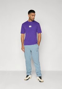 Vijolična bombažna majica s belim logotipom na prsih, kombinirana s svetlo modrimi trenirkami z majhnim črnim logotipom in črno-belimi supergami.