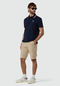 Hombre con polo azul oscuro con ribete naranja, pantalones cortos cargo beige, zapatillas blancas y gafas de sol negras, de pie con las manos en los bolsillos.