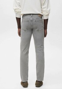 Persona con jeans rectos gris claro, top blanco de manga larga y zapatos marrones, de pie de espaldas frente a un fondo liso.