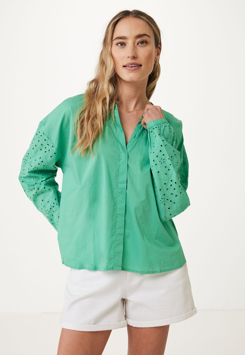 Camicia verde con maniche lunghe e ricami ad occhio, abbinata a pantaloni corti bianchi. Il tessuto ha una texture leggera e ariosa.
