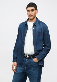 Τζιν πουκάμισο σε σκούρο μπλε χρώμα με κουμπιά μπροστά, φορεμένο πάνω από ένα λευκό μπλουζάκι. Διαθέτει ένα μικρό κόκκινο λογότυπο και μια ασημένια αλυσίδα στο λαιμό.