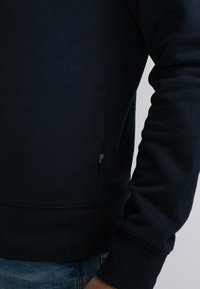 Granatowy sweatshirt wykonany z miękkiej bawełny, z ribbowanymi mankietami i dolnym wykończeniem. Widok z boku podkreśla małą metkę marki przy szwie.