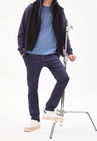 Muž v modrých kalhotách, modré košili, černé mikině a bílých teniskách stojí s jednou nohou na kovovém podstavci na bílém pozadí.