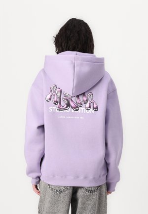 Personne portant un sweat à capuche lavande avec un logo "ALPHA" rose brillant et un texte blanc dans le dos, associé à un jean gris.