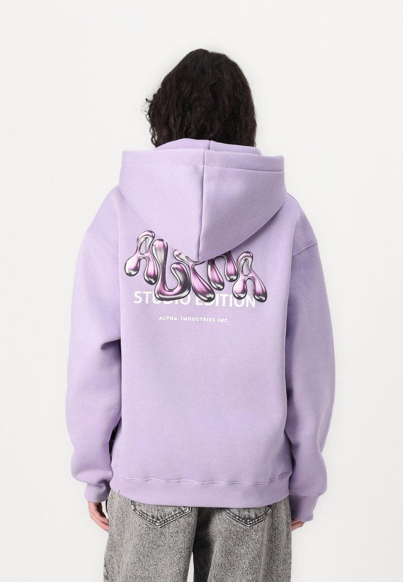 Personne portant un sweat à capuche lavande avec un logo "ALPHA" rose brillant et un texte blanc dans le dos, associé à un jean gris.
