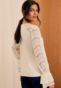 Weiße Bluse mit langen Spitzenärmeln, die ein florales Muster und Rüschensäume aufweisen. Der Körper hat eine glatte Textur und eine lockere Passform.