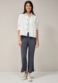 Witte denim jacket met knoopsluiting, gecombineerd met losse donkergrijze broek. Model draagt witte sneakers en staat tegen een neutrale achtergrond.