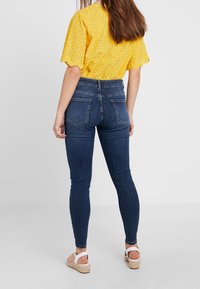 Höga, mörkblå jeans med stretchig passform, har två bakfickor och detaljerad sömnad. Bärs med en gul texturerad topp och sandaler.