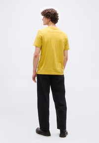 Jeune homme aux cheveux bouclés, debout de dos, portant une chemise jaune moutarde à manches courtes, un pantalon noir et des chaussures noires.