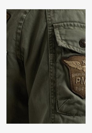 Olijfgroene jacket met een stoffen patch met geborduurde details, een klepzak en een metalen knoopaccent op de schouder.