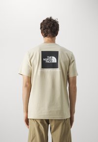 T-shirt en coton beige avec un graphique carré noir portant le logo "THE NORTH FACE" et le texte "NEVER STOP EXPLORING" au dos.