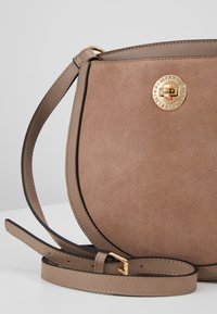 Borsa rotonda in suede color sabbia con una chiusura a logo dorato, caratterizzata da una tracolla in pelle liscia e da un bordo in contrasto nero lungo i lati.
