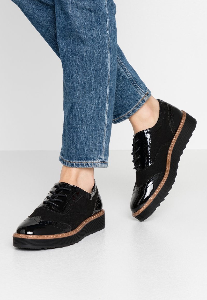 Chaussures brogue noires en cuir poli et en daim avec des semelles épaisses, portées avec un jean en denim bleu sur un sol blanc.