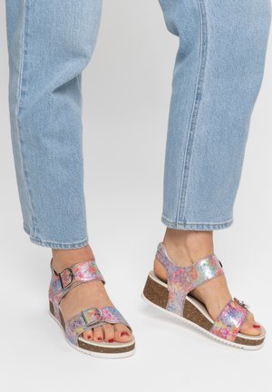 Feet wearing light blue jeans and colorful floral-patterned wedge sandals with buckles, standing on a white surface.