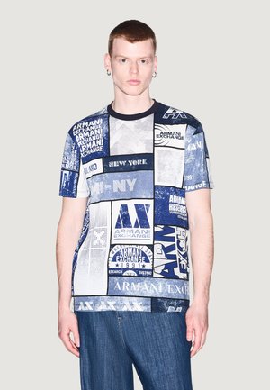 Giovane uomo con capelli rossi corti indossa una maglietta patchwork blu e bianca Armani Exchange e jeans blu su uno sfondo chiaro e uniforme.
